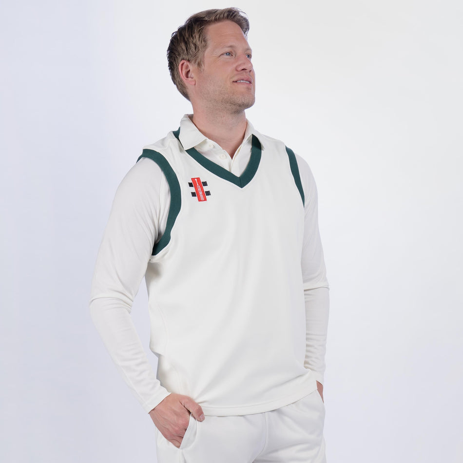 A man wears a Gray Nicolls Oldlands CC Pro Performance Slipover over a white cricket uniform, hands in pockets, looking upward and smiling against a plain light background.
