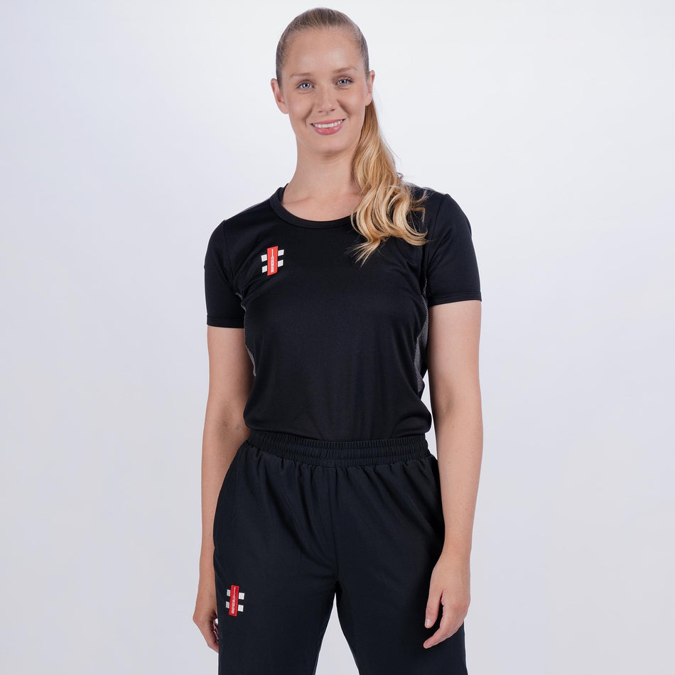 A woman with blonde hair in a ponytail wears the Gray Nicolls Pro Performance Ladies T-Shirt in black, featuring small red and white brand logos—ideal for moisture wicking during high intensity training against a light background.
