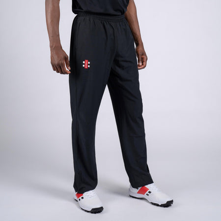 A person wears Gray Nicolls Rockhampton CC Velocity Track Trouser - Junior in black with a small red and white logo on the left thigh, paired with white sneakers with red and black accents, standing against a plain light background.