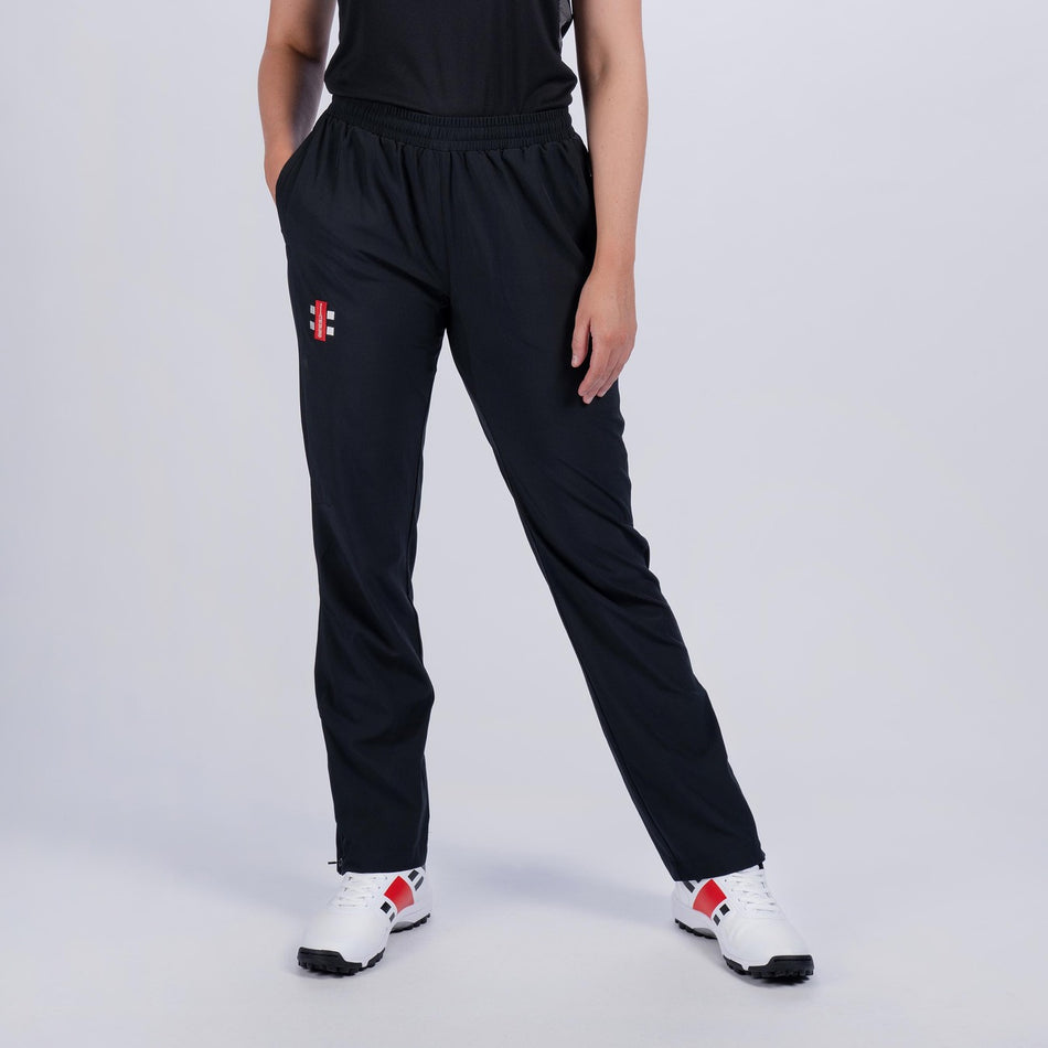 A woman wearing Gray Nicolls Velocity Ladies Track Trousers, featuring zipped pockets and a red and white logo, pairs them with a black shirt and white sneakers with red and black details while standing against a plain light background.