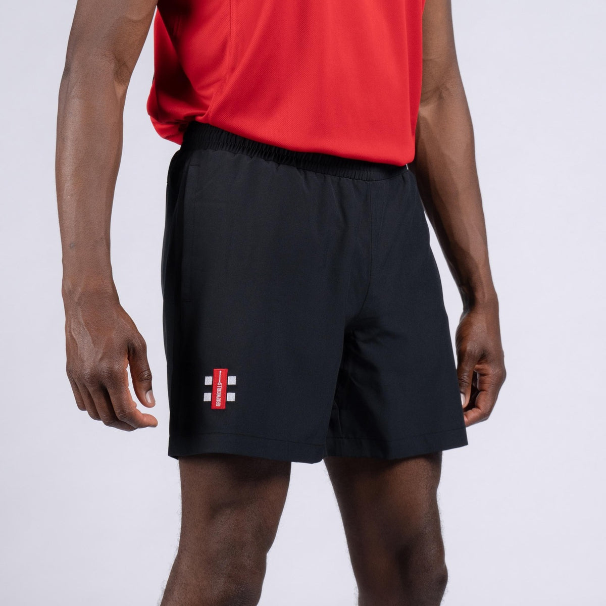 A person in a red shirt and black Gray Nicolls Rockhampton CC Velocity Shorts - Junior, featuring a small logo on the left leg, stands against a plain light background.