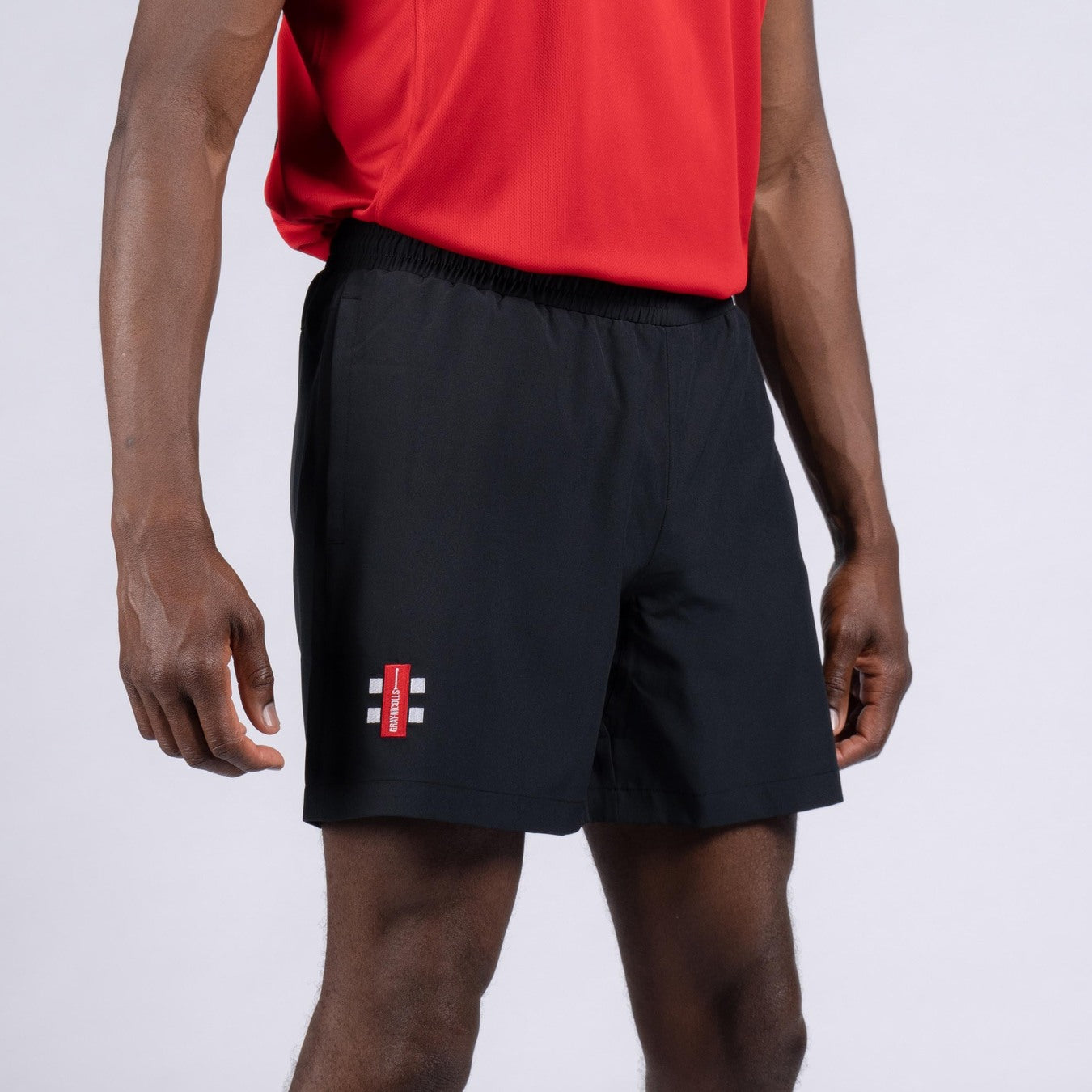 A person in a red shirt and black Gray Nicolls Rockhampton CC Velocity Shorts - Junior, featuring a small logo on the left leg, stands against a plain light background.