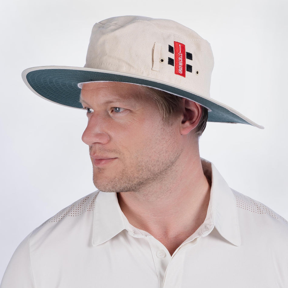 A man in a white collared shirt looks left against a plain background, wearing a light-colored Gray Nicolls Rockhampton CC Sunhat with a red tag and black ventilation holes.