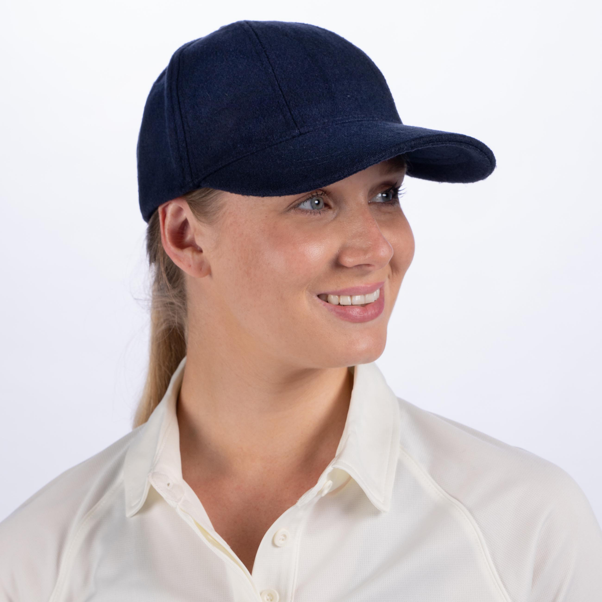 A woman in a Gray Nicolls Monmouth CC Melton County Cap with a navy club logo and a cream collared shirt smiles and looks to the side against a plain white background.