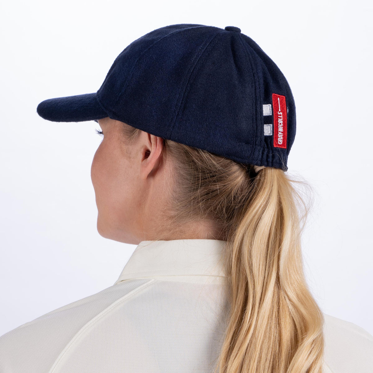 A woman with long blonde hair in a ponytail is seen from behind, wearing a Gray Nicolls Monmouth CC Melton County Cap with a red and white patch and a cream collared shirt, set against a plain white background.