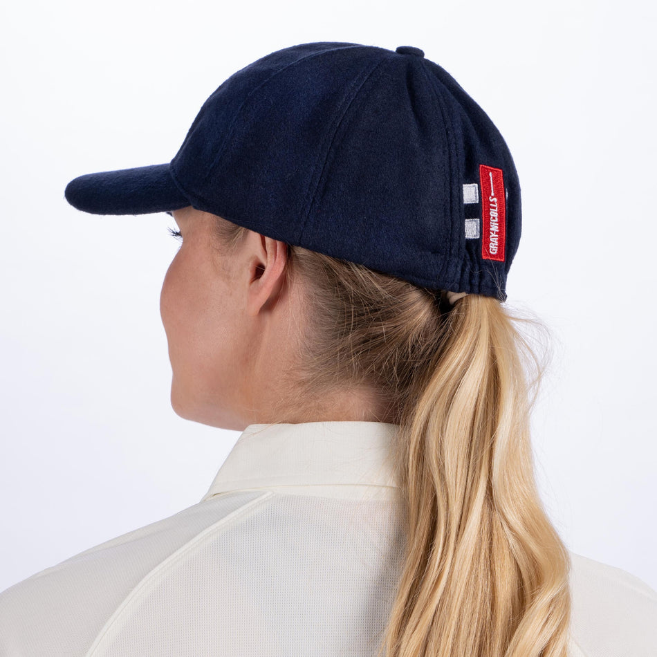 A woman with long blonde hair in a ponytail is seen from behind, wearing a Gray Nicolls Monmouth CC Melton County Cap with a red and white patch and a cream collared shirt, set against a plain white background.