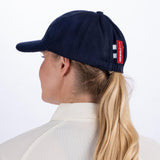 A woman with blonde hair in a ponytail, shown from behind, wears a Gray Nicolls Melton County Cap in navy (senior size) and a light collared shirt. The traditional style cap features a red tag with white text on the adjustable strap.