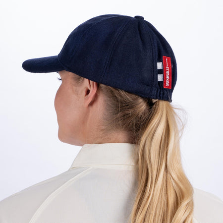 A woman with blonde hair in a ponytail, shown from behind, wears a Gray Nicolls Melton County Cap in navy (senior size) and a light collared shirt. The traditional style cap features a red tag with white text on the adjustable strap.