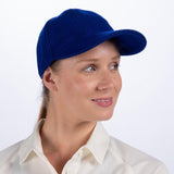 A woman wearing the Gray Nicolls Melton County Cap in royal by Gray Nicolls and a white collared shirt smiles slightly to the side against a plain light background.