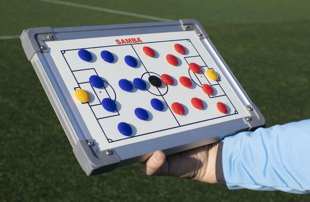 A coach uses the Samba Sports Mini Tactic Board with red, blue, yellow, and black magnetic markers on a soccer field diagram outdoors. A wipe-board marker is included for quick strategy changes on the pitch.