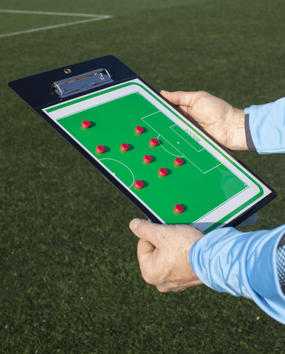 A person holds a Samba Sports Samba Soccer Coaches Clipboard with a tactics board displaying red markers on a green field, standing on grass near a soccer pitch.