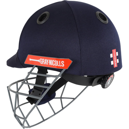 The Gray Nicolls Atomic Helmet is a lightweight junior cricket helmet in navy blue, featuring a metal faceguard, black ventilation holes, red side logo, and an adjustable back strap for a secure fit.