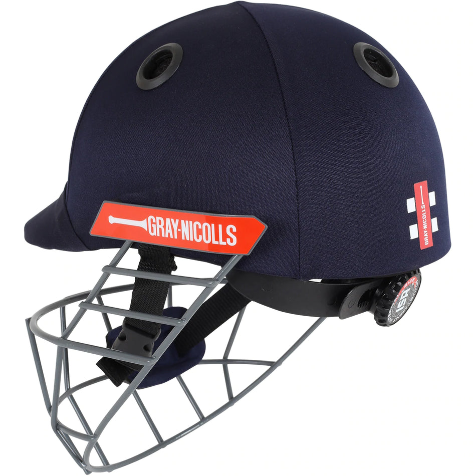 The Gray Nicolls Atomic Helmet is a lightweight junior cricket helmet in navy blue, featuring a metal faceguard, black ventilation holes, red side logo, and an adjustable back strap for a secure fit.