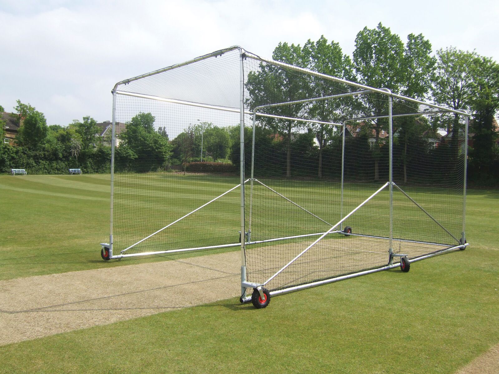 The Harrod Sport Premier Wheelaway Cricket Cage, made of galvanised steel, stands on a grass field beside a cricket pitch, with mesh sides and a sturdy frame visible among trees and houses in the background.