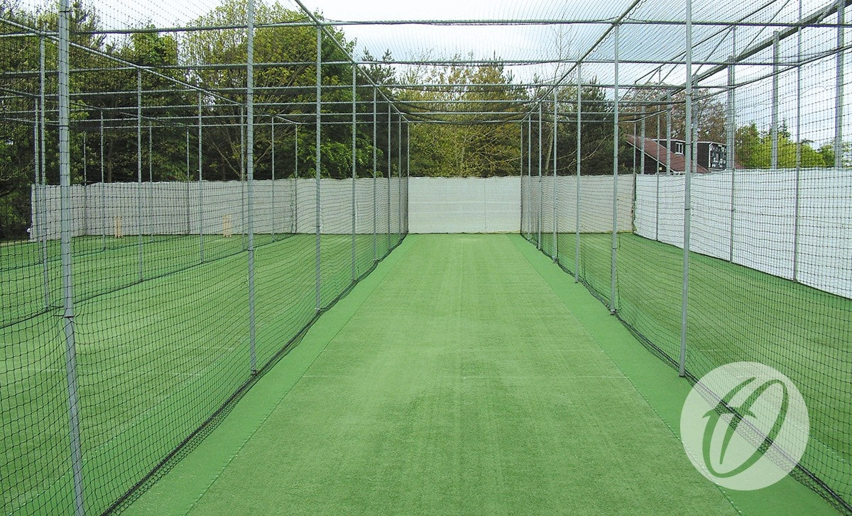 The Harrod Sport Heavy Duty Cage - Sockets is a green outdoor cricket practice net with synthetic turf, enclosed by sturdy cages and white fencing, with trees visible in the background.