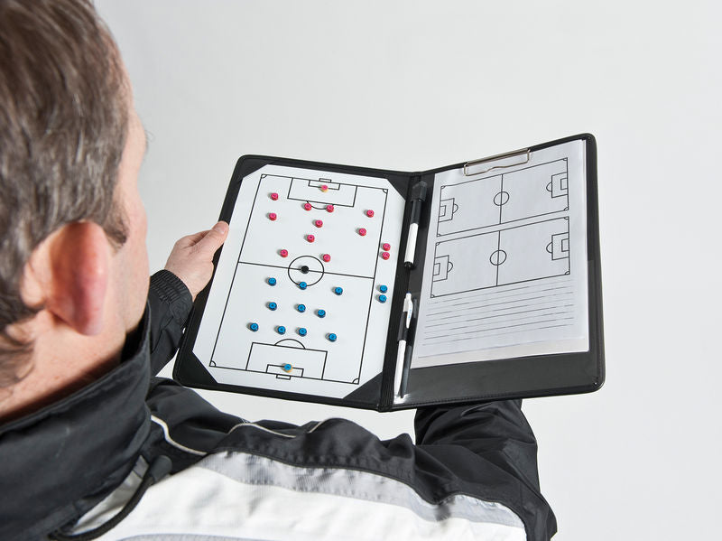 A coach uses the Precision Training Precision Pro Soccer Coaches Tactic Folder, displaying a soccer field diagram with red and blue magnetic markers to plan strategy. The open folder shows a dry-erase marker and notes section for tactical planning.