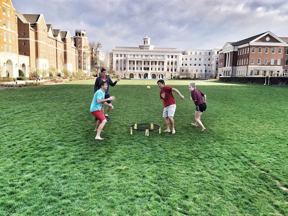 Four people play barefoot on a grassy field, enjoying the Kankakee Spikeball Standard Set with its adjustable net. This lively outdoor game brings friendly competition under a cloudy sky, surrounded by brick and white buildings.