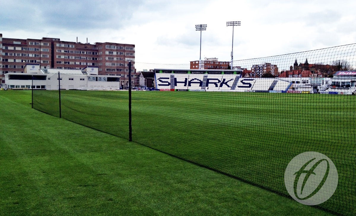 A cricket field featuring the Harrod Sport County Practice Net System - 40M with polypropylene netting supported by sturdy aluminium uprights, surrounded by green grass, stadium seating labeled SHARKS, and buildings in the background beneath a cloudy sky.