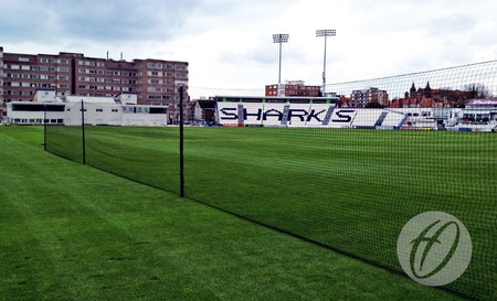 A cricket field featuring the Harrod Sport County Practice Net System - 40M with polypropylene netting supported by sturdy aluminium uprights, surrounded by green grass, stadium seating labeled SHARKS, and buildings in the background beneath a cloudy sky.