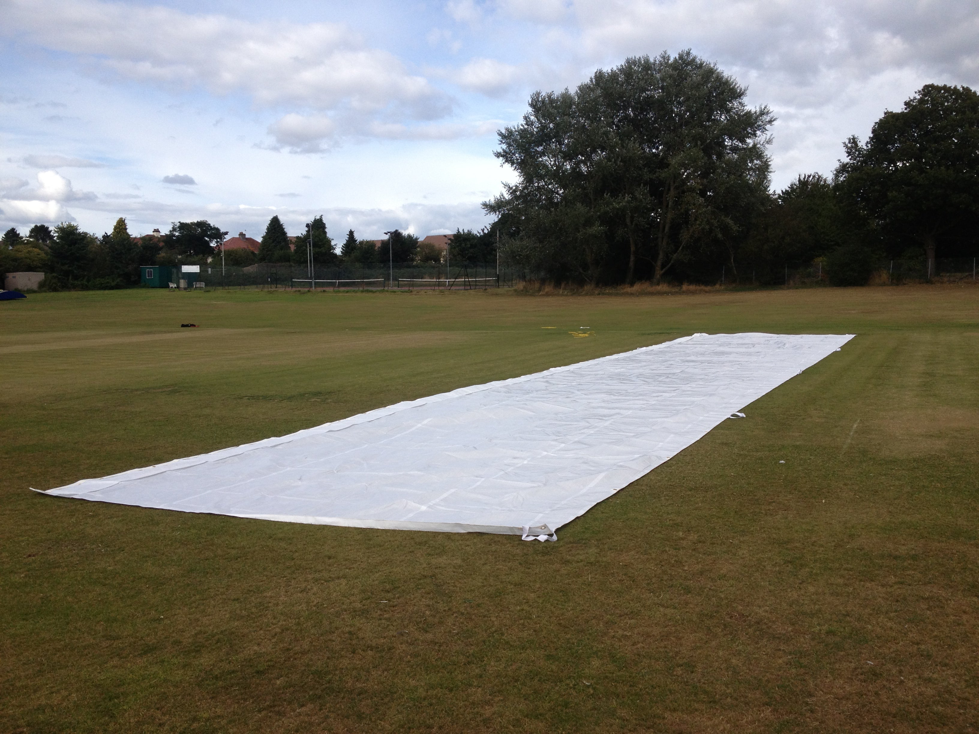 A Stuart Canvas Flatlay Lightweight Pitch Cover (25m x 6m) is spread out on a grassy field, with trees, fencing, and buildings visible in the background beneath a partly cloudy sky.
