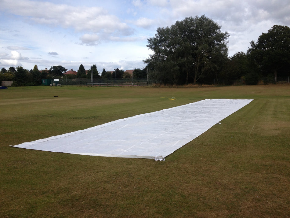 A Stuart Canvas Flatlay Lightweight Pitch Cover (25m x 6m) is spread out on a grassy field, with trees, fencing, and buildings visible in the background beneath a partly cloudy sky.