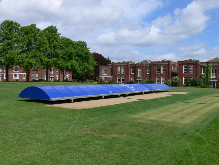 The Ultimate Mobile Cricket Pitch Covers by Martin Berrill Sports protect the field’s center, surrounded by green grass and red-brick buildings under a partly cloudy sky, demonstrating the product's reliable performance.