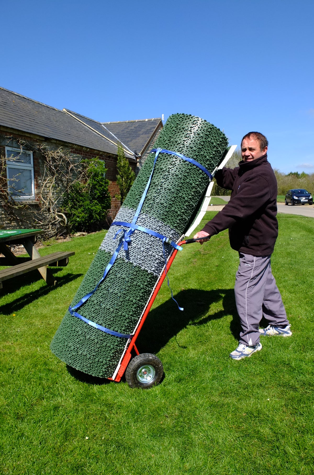 A man stands on grass, using a Flicx UK Flicx Safety Trolley to transport large rolls of green 2G Flicx Pitch outside a brick house on a sunny day—demonstrating easy pitch movement.