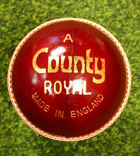 A red Dukes County Royal Cricket Ball Senior, made from English alum tanned leather and featuring gold "A County Royal Made in England" text, lies on a green textured surface with prominent seams and stitching.