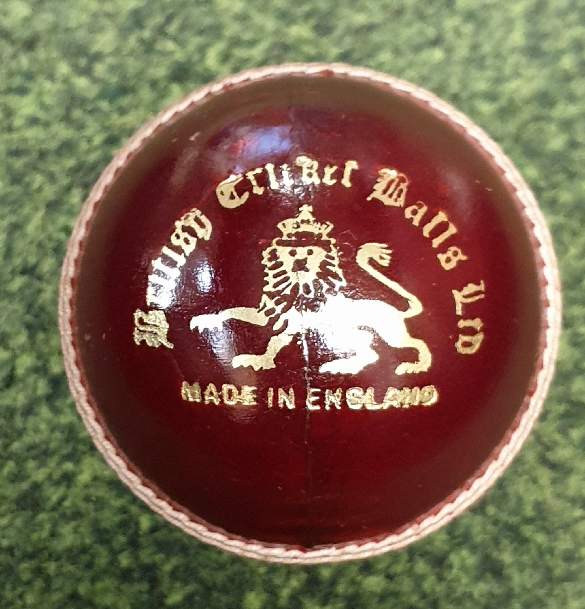 A close-up of the Dukes County Royal Cricket Ball Senior, made from English alum tanned leather with gold embossing, including a lion emblem, rests on a green textured surface. The ball is crafted by Dukes in England.