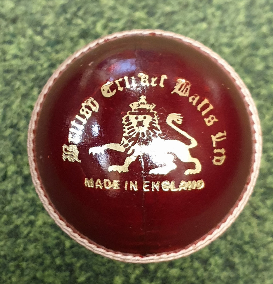 A close-up of the Dukes County Royal Cricket Ball Senior, made from English alum tanned leather with gold embossing, including a lion emblem, rests on a green textured surface. The ball is crafted by Dukes in England.