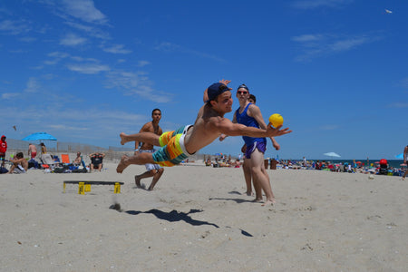 A young man dives on a sandy beach to hit a yellow ball from the Kankakee Spikeball Standard Set, enjoying this lively game with friends as others relax under umbrellas on a sunny day.