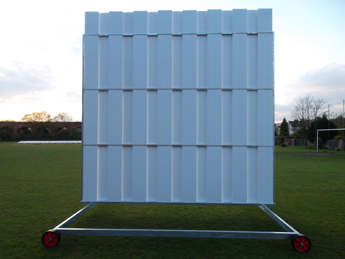 The Stuart Canvas Polypropylene Sightscreen, large and white with wheels for mobility, stands on a grassy field beneath a partly cloudy sky and surrounded by trees.