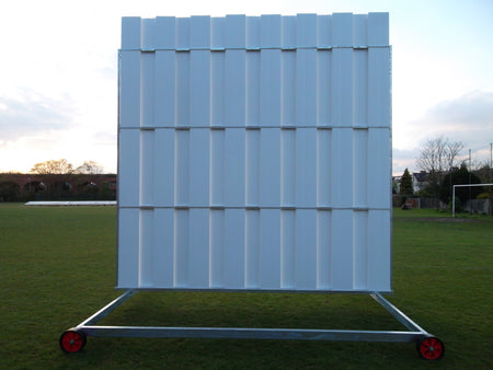 The Stuart Canvas Polypropylene Sightscreen, large and white with wheels for mobility, stands on a grassy field beneath a partly cloudy sky and surrounded by trees.