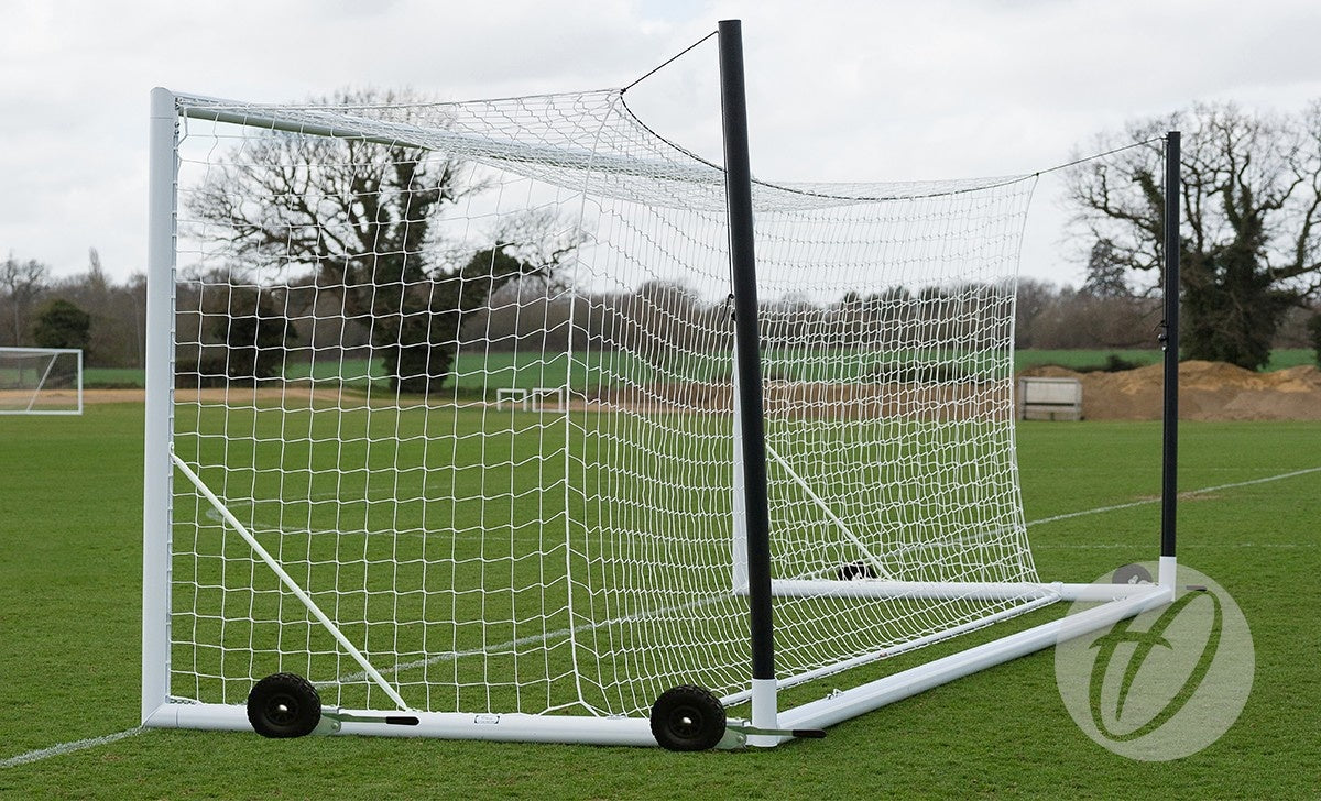 The Harrod Sport 4G Euro Portagoal - 7V7/5V5 is positioned on a grassy field. Its net is secured to a reinforced white aluminium frame, and two black support poles extend backward. Trees and cloudy skies form the background.
