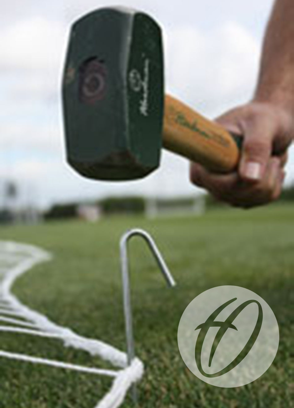 A person uses a mallet to drive Harrod Sport 180MM Steel Ground Pegs into grass, securing a white boundary line on a sports field. A circular watermark with a stylized H logo appears in the bottom right corner.