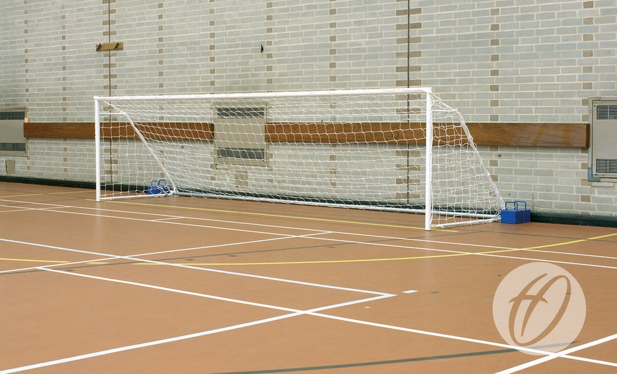 A Harrod Sport FS1 Fold-away Goal (4.88m x 1.22m, 5-a-side) stands on a brown gym floor with white court lines near a brick wall, its frame secured in place with blue weights.