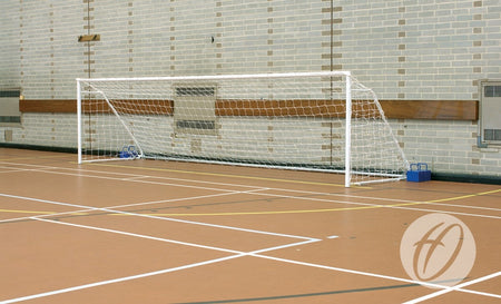 A Harrod Sport FS1 Fold-away Goal (4.88m x 1.22m, 5-a-side) stands on a brown gym floor with white court lines near a brick wall, its frame secured in place with blue weights.