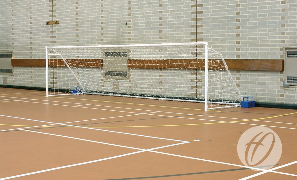 A Harrod Sport FS1 Fold-away Goal (4.88m x 1.22m, 5-a-side) stands on a brown gym floor with white court lines near a brick wall, its frame secured in place with blue weights.