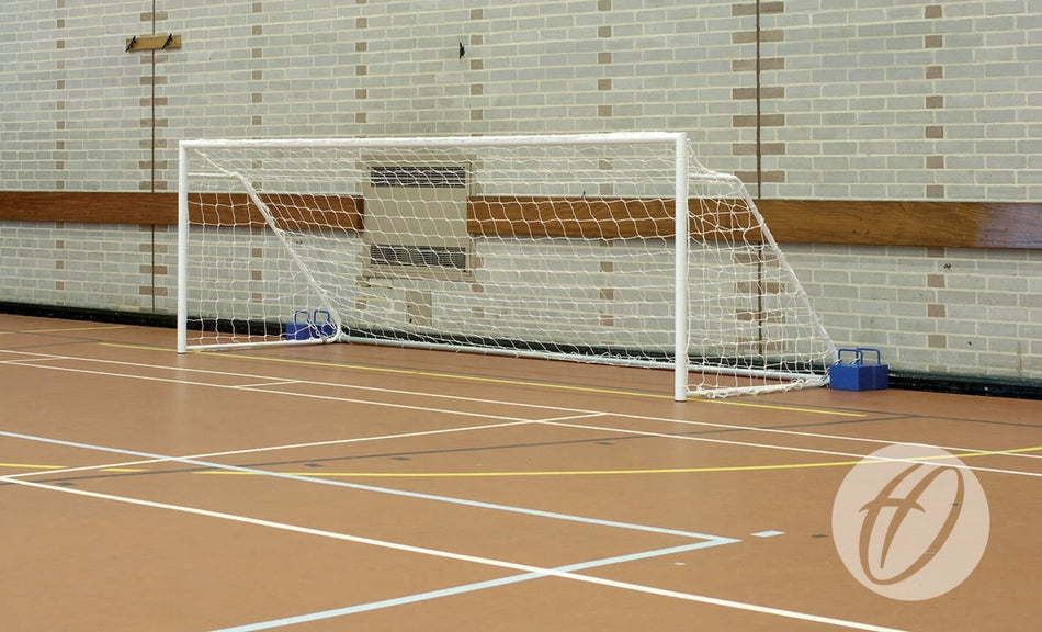 An indoor sports hall featuring tan flooring and white court lines is equipped with the Harrod Sport FS1 Fold-away Goal – 3.66m x 1.22m for 5-a-side play, set against a brick wall. This sturdy steel goal has a white frame with netting and is securely anchored at the posts by two blue weights.