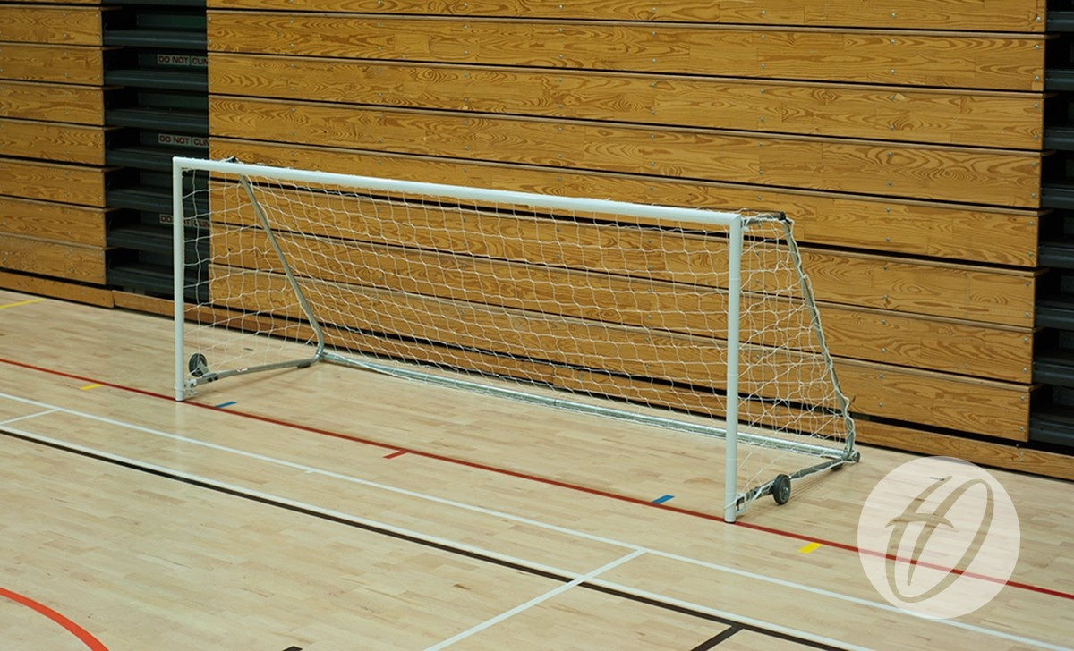 A Harrod Sport FS1 Fold-away Goal (2.44m x 1.22m, 5-a-side) with a net is set up on a light-colored indoor gym floor in a sports hall, positioned in front of a wooden wall featuring horizontal panels and black accents.