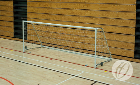 A Harrod Sport FS1 Fold-away Goal (2.44m x 1.22m, 5-a-side) with a net is set up on a light-colored indoor gym floor in a sports hall, positioned in front of a wooden wall featuring horizontal panels and black accents.
