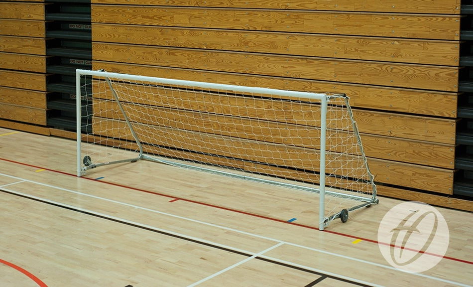 A Harrod Sport FS1 Fold-away Goal (2.44m x 1.22m, 5-a-side) with a net is set up on a light-colored indoor gym floor in a sports hall, positioned in front of a wooden wall featuring horizontal panels and black accents.