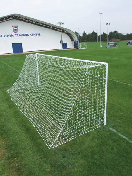 A Harrod Sport Heavyweight Football Goal - Senior with white steel frame and netting stands on a neatly cut green field; in the background, a building labeled TRAINING CENTRE is visible along with additional junior club football goals. The sky is cloudy.