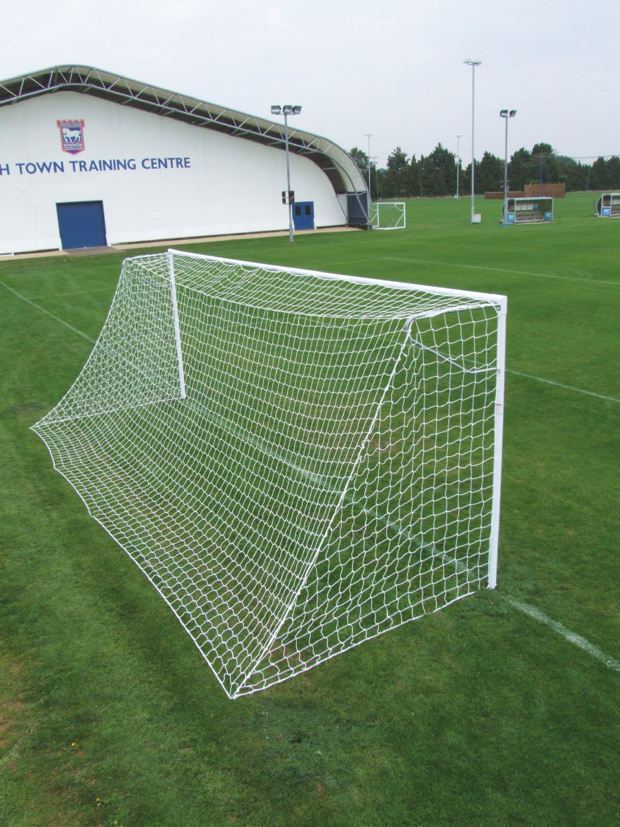 A Harrod Sport Heavyweight Football Goal - Senior with white steel frame and netting stands on a neatly cut green field; in the background, a building labeled TRAINING CENTRE is visible along with additional junior club football goals. The sky is cloudy.