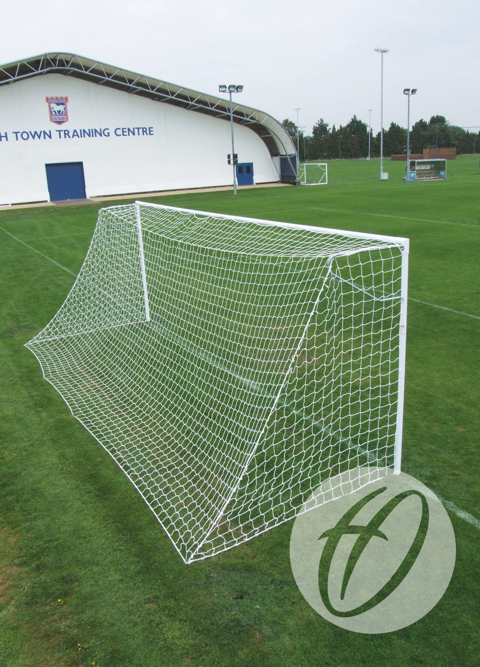 A Harrod Sport Heavyweight Football Goal – Junior, crafted from white steel and fitted with netting, stands on a green grass field near a building labeled TOWN TRAINING CENTRE under a clear sky—making it ideal as a sturdy, socketed football goal for regular junior club training.