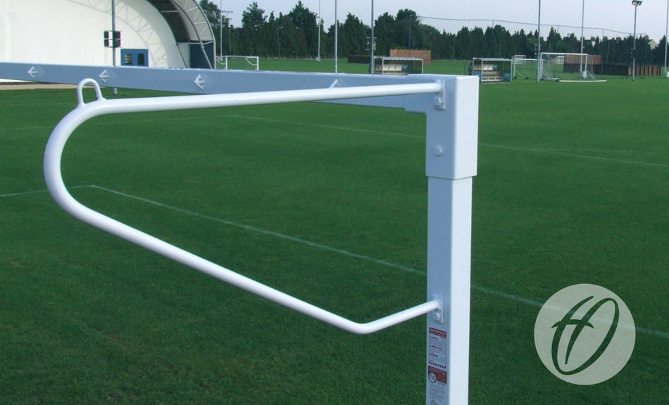 A white metal gate with a curved bar is installed on the edge of a green soccer field, accompanied by Harrod Sport Solid Steel Continental Net Supports - Senior/Junior and anti-vandal goals, with trees visible in the background.