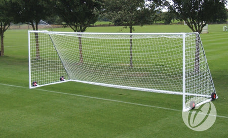 A Harrod Sport Heavyweight Freestanding Football Goal – Senior, featuring a white, fully welded crossbar and red wheels, with an attached net, stands on a green grass field surrounded by trees in the background.