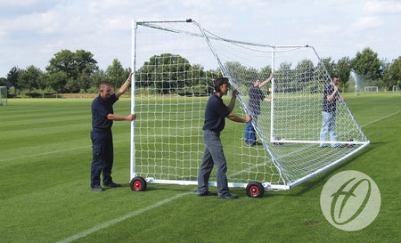 Four people are moving a Harrod Sport Heavyweight Freestanding Football Goal – Senior, which is equipped with wheels, across a grassy field. The weather is clear, and trees as well as additional soccer goals are visible in the background. This robust steel goal from Harrod Sport features a fully welded crossbar for enhanced durability and easy mobility, making it suitable for players of all ages.