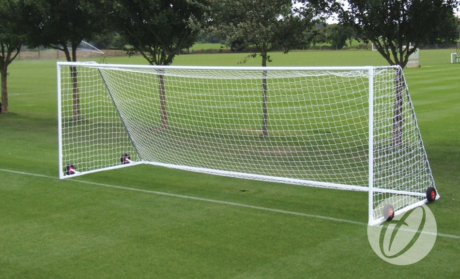 A Harrod Sport Heavyweight Freestanding Football Goal - Junior, featuring a white steel frame and netting, stands on green grass with trees and a field in the background. This junior-sized goal is equipped with four small red wheels at its base for easy maneuverability and is designed to be safe for use on artificial surfaces.