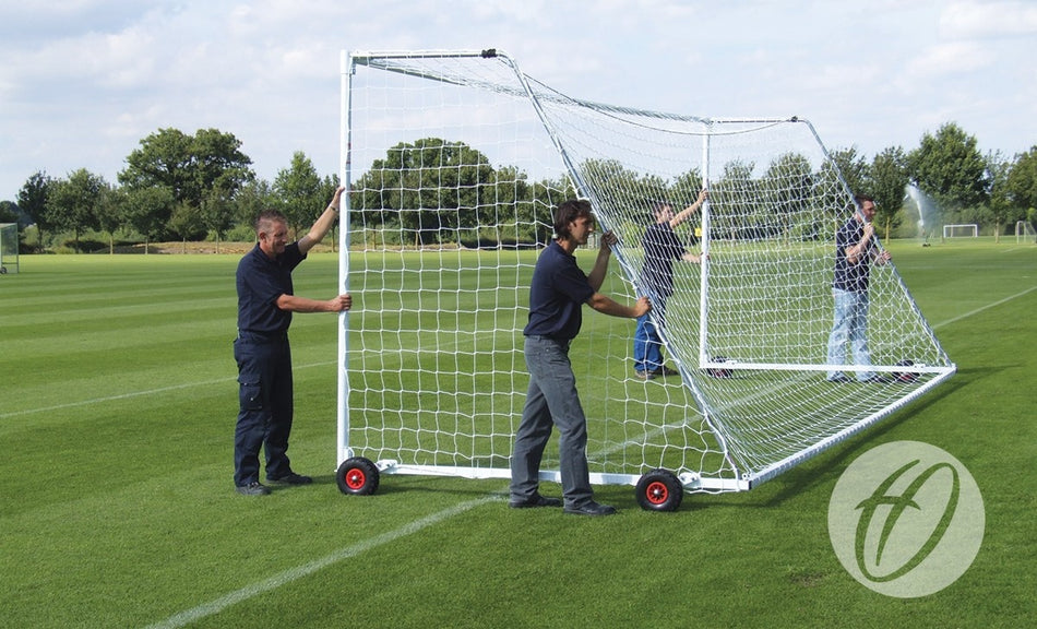Four people are moving a Harrod Sport Heavyweight Freestanding Football Goal - Junior across a grassy field using the wheels attached to its frame. Trees and additional freestanding football goals can be seen in the background under a partly cloudy sky.